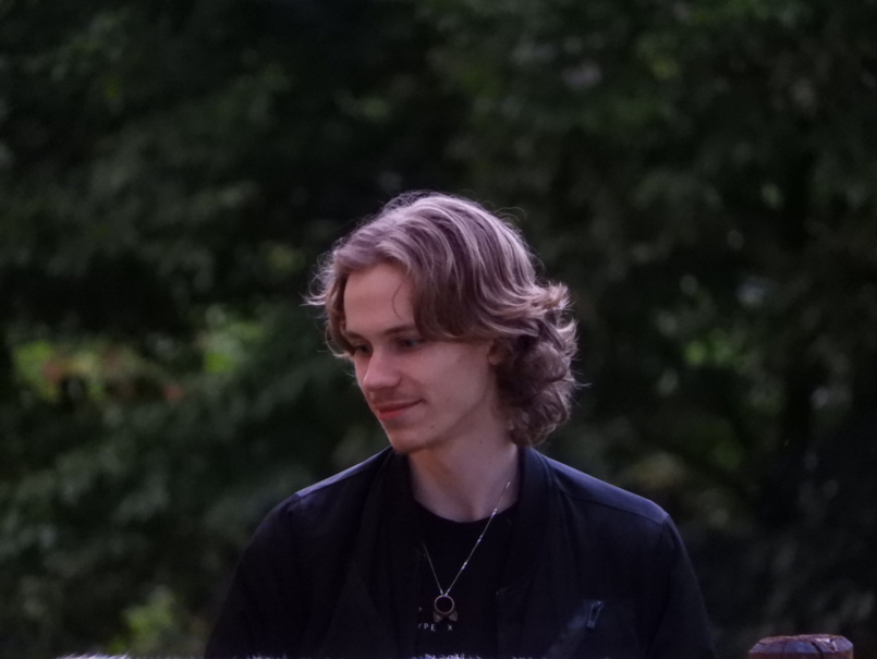 Photo of Kjell, standing in a grass field, in the background green leaves of distant trees. He has a mild smile on his face, his head is tilted towards the ground ever so slightly.